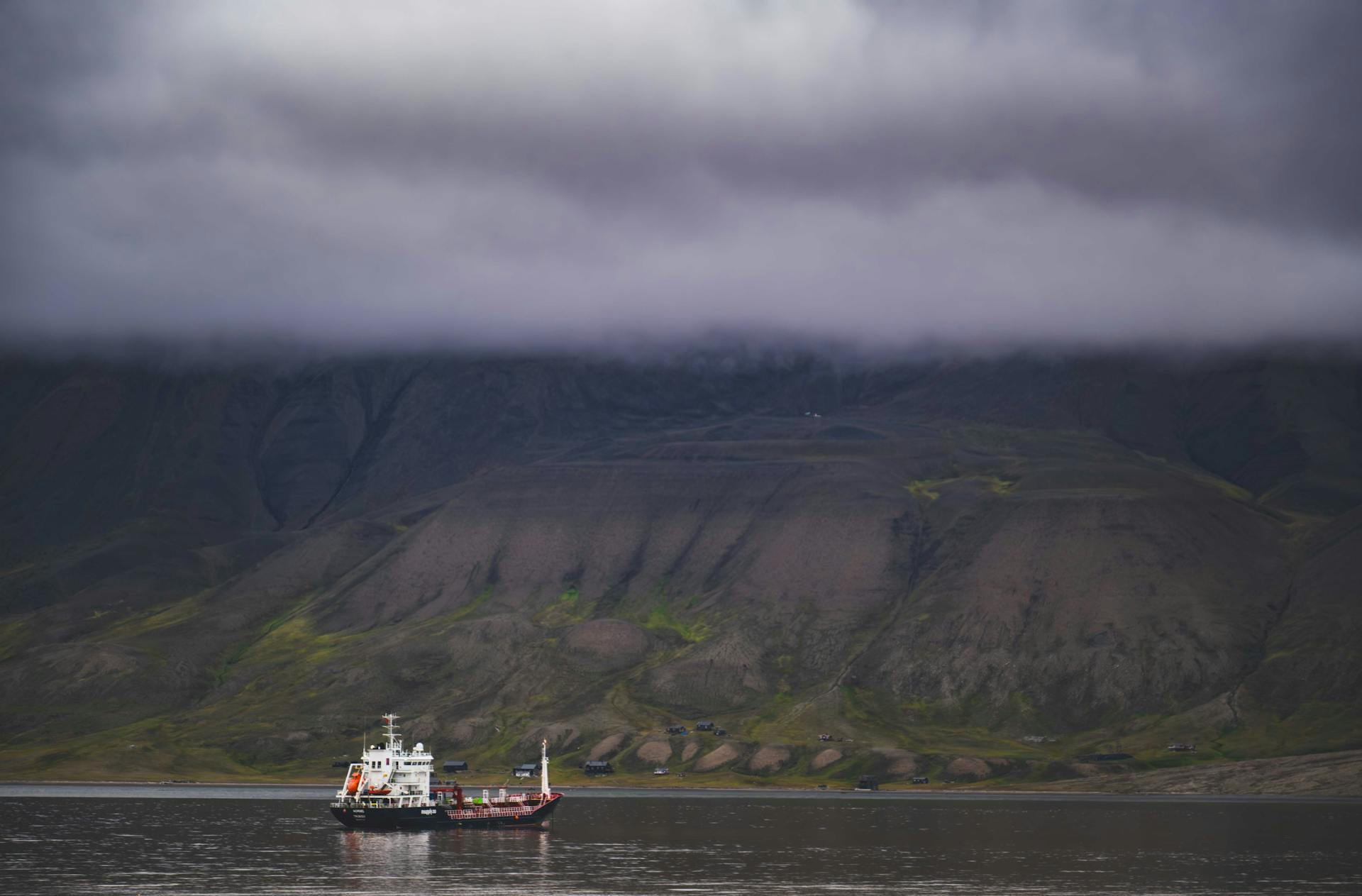 A scenic view of a tanker ship sailing in front of dramatic mountains under cloudy skies.