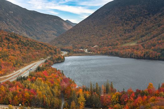 Aerial view of a vibrant autumn landscape with a winding road by a clear lake and forested hills.