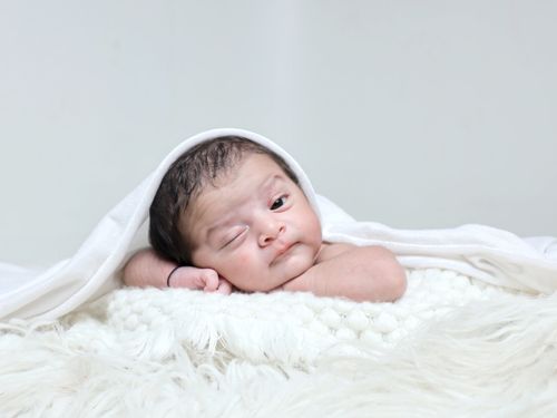 Free Adorable newborn baby with black hair winking, wrapped in a soft white blanket. Stock Photo