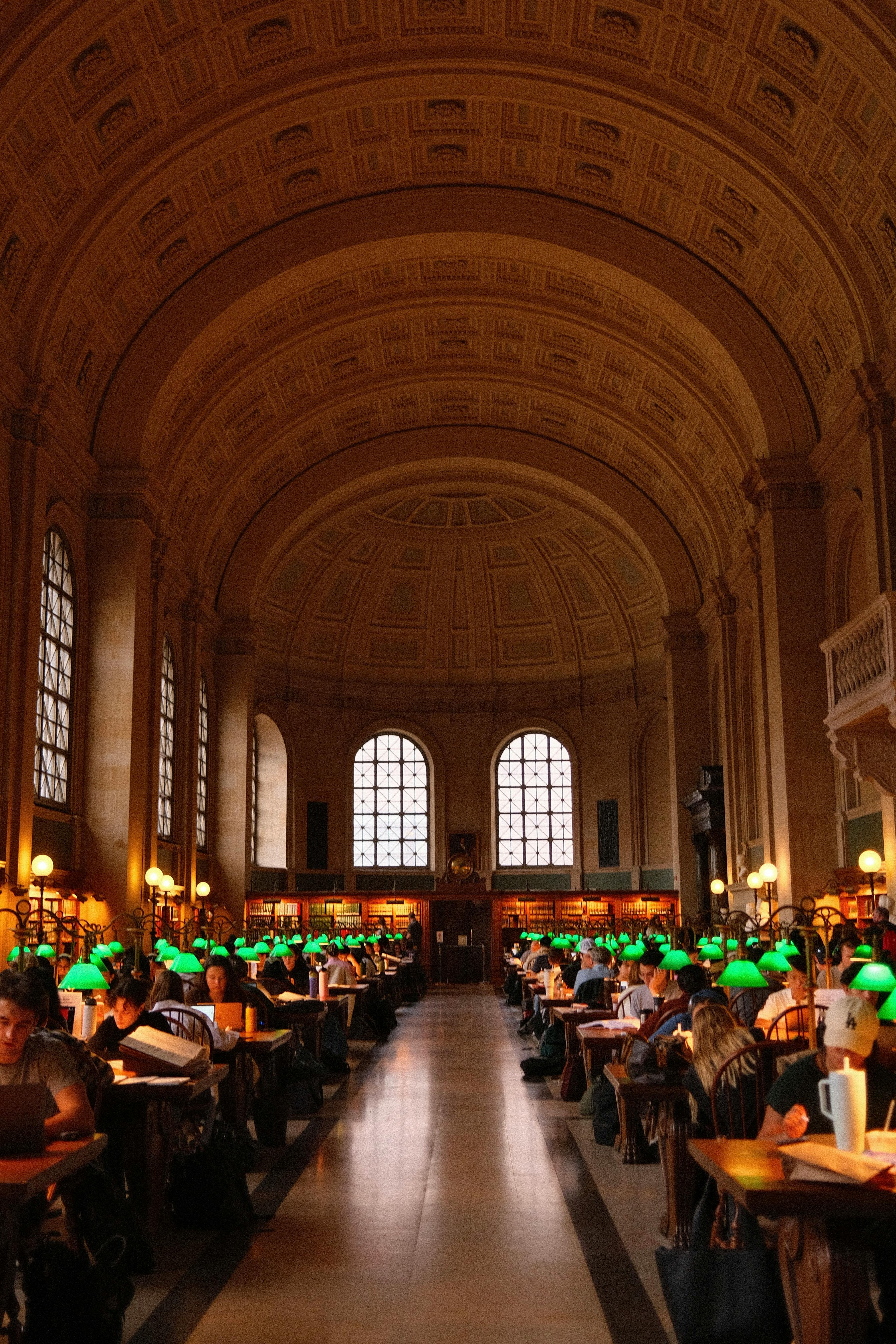 People studying in the grand reading room of Boston Public Library with iconic arched ceiling and green lamps.