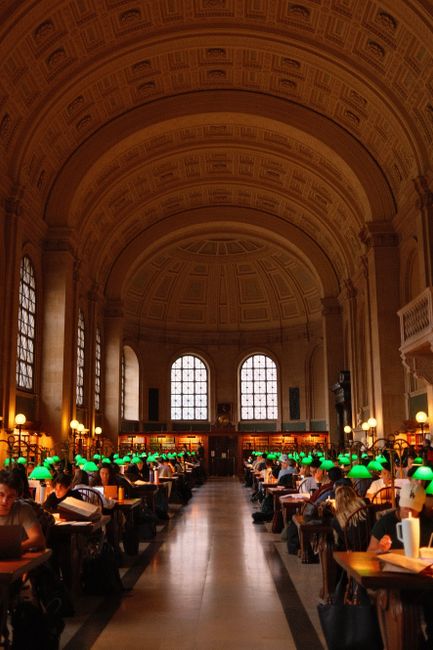 People studying in the grand reading room of Boston Public Library with iconic arched ceiling and green lamps.