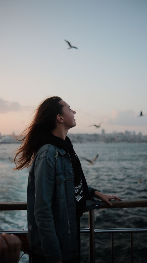 Free A woman stands by the sea at dusk, enjoying the view of birds and city skyline. Stock Photo