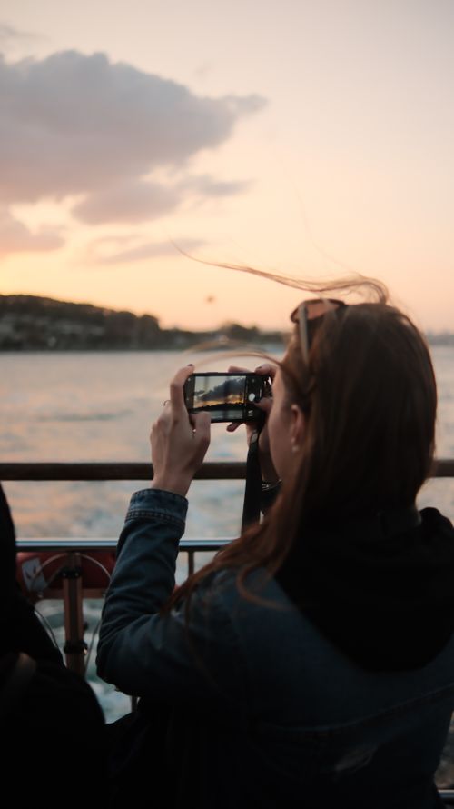 Free A woman with long brown hair takes a photo of the sunset from a boat using her smartphone. Stock Photo