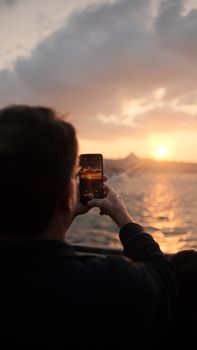 A woman photographs the scenic Istanbul sunset by the sea, emphasizing travel and technology.