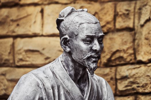 A detailed bronze sculpture of a historical figure set against a textured brick wall background.