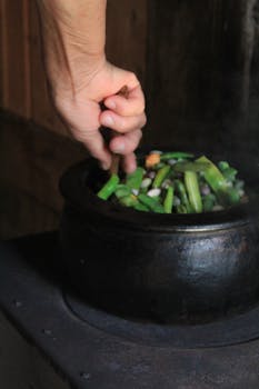 Close-up of a hand stirring fresh vegetables in a black pot, capturing traditional cooking at its finest.