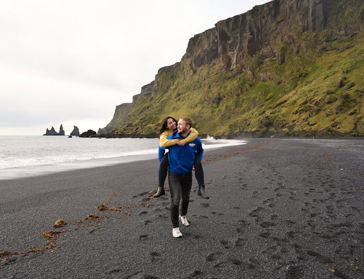 A Couple Walking On A Black Beach With Mountains In The Background