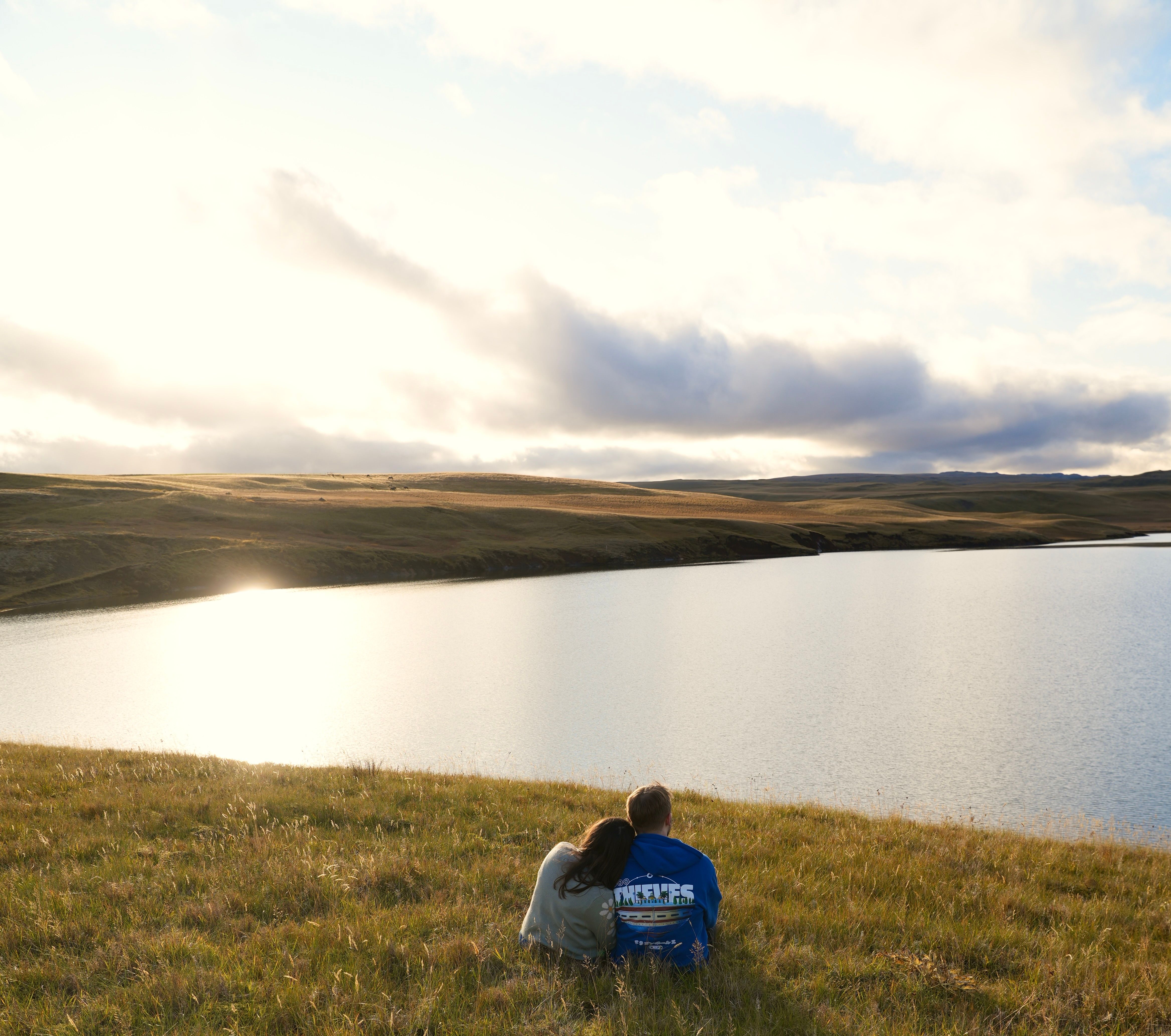 Two people sitting on a grassy hill overlooking a lake · Free Stock Photo