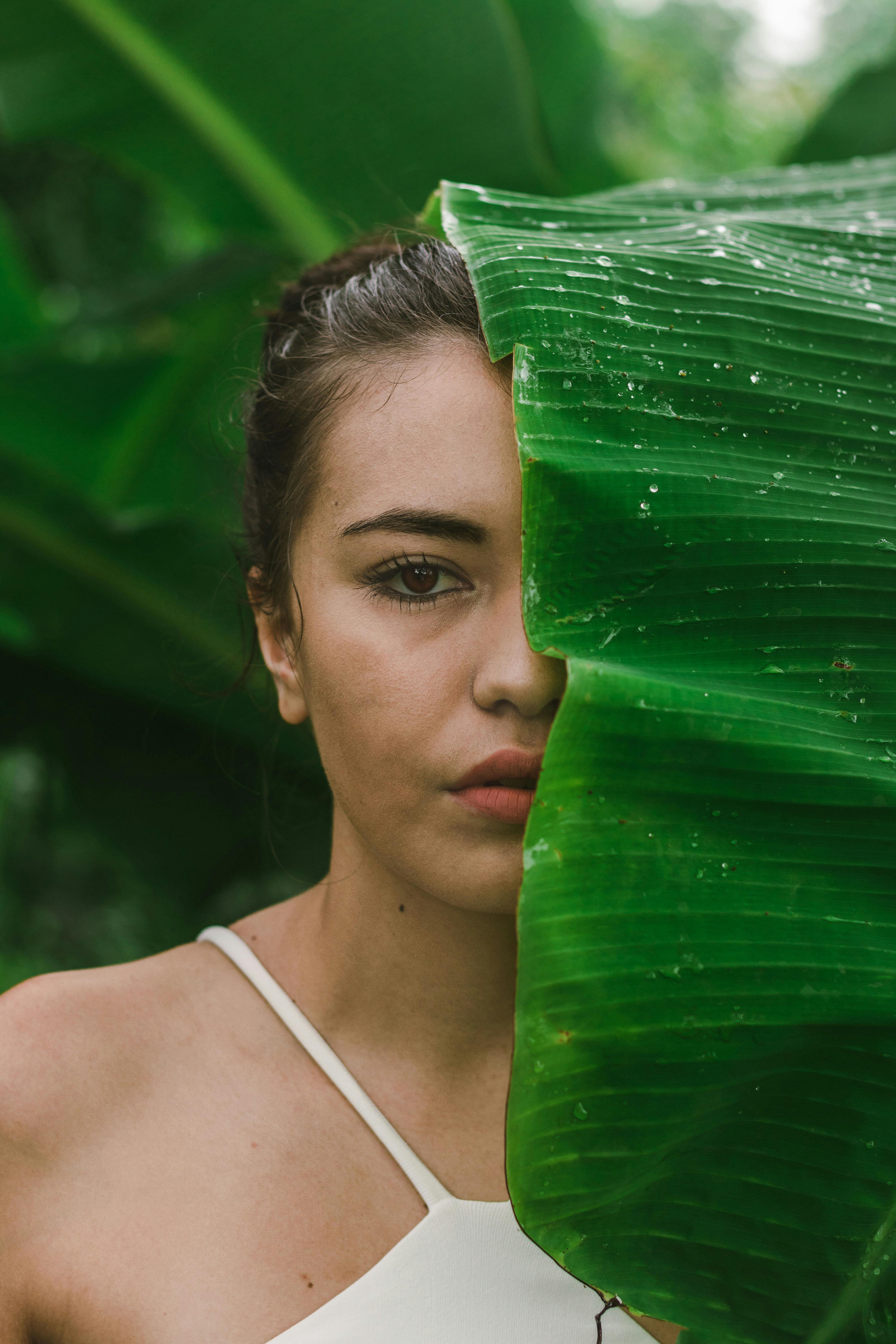 Portrait of a woman partially hidden behind a vibrant banana leaf outdoors.