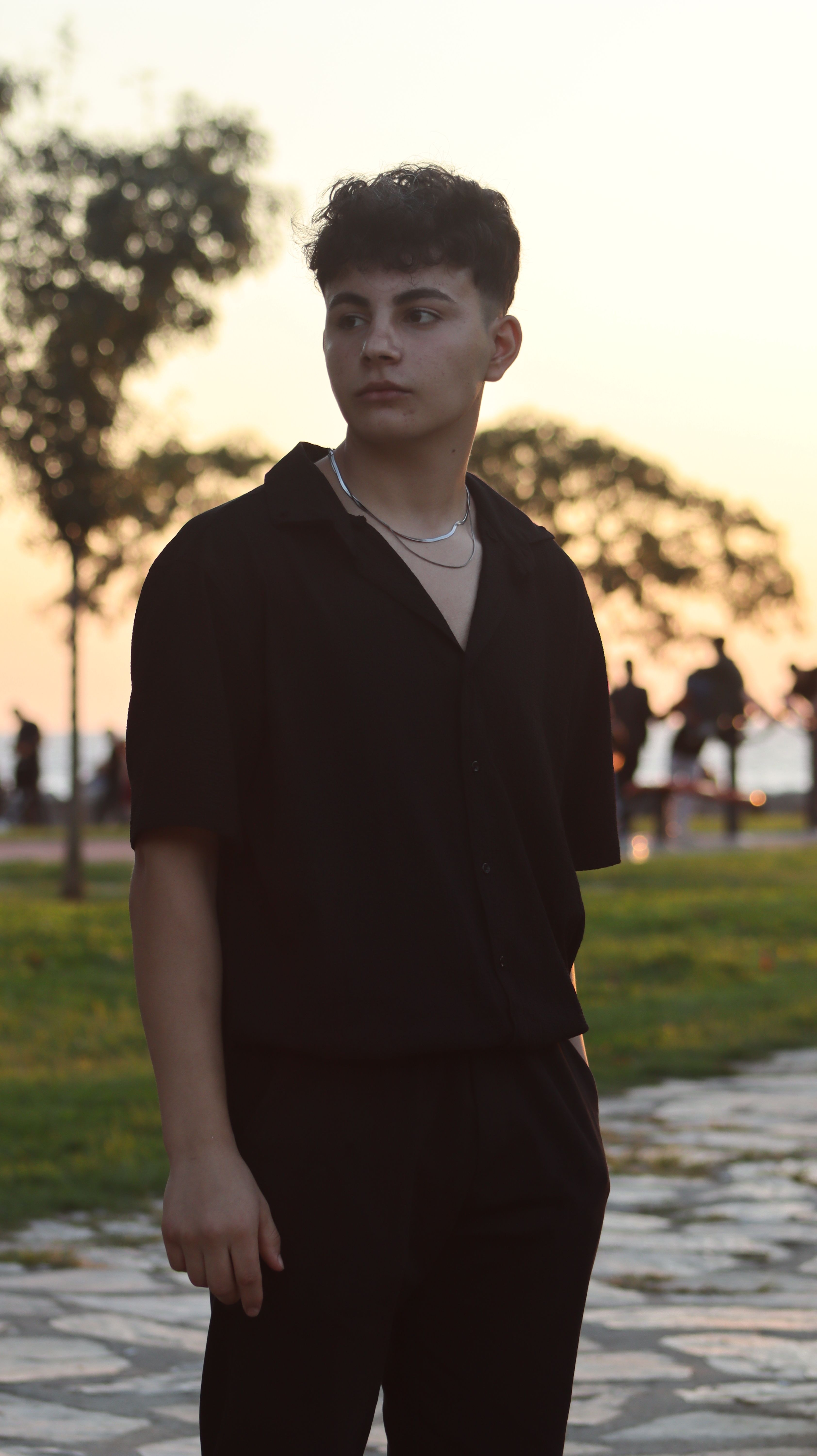 Portrait of a young man in a black shirt standing in a park at sunset, showcasing urban fashion.