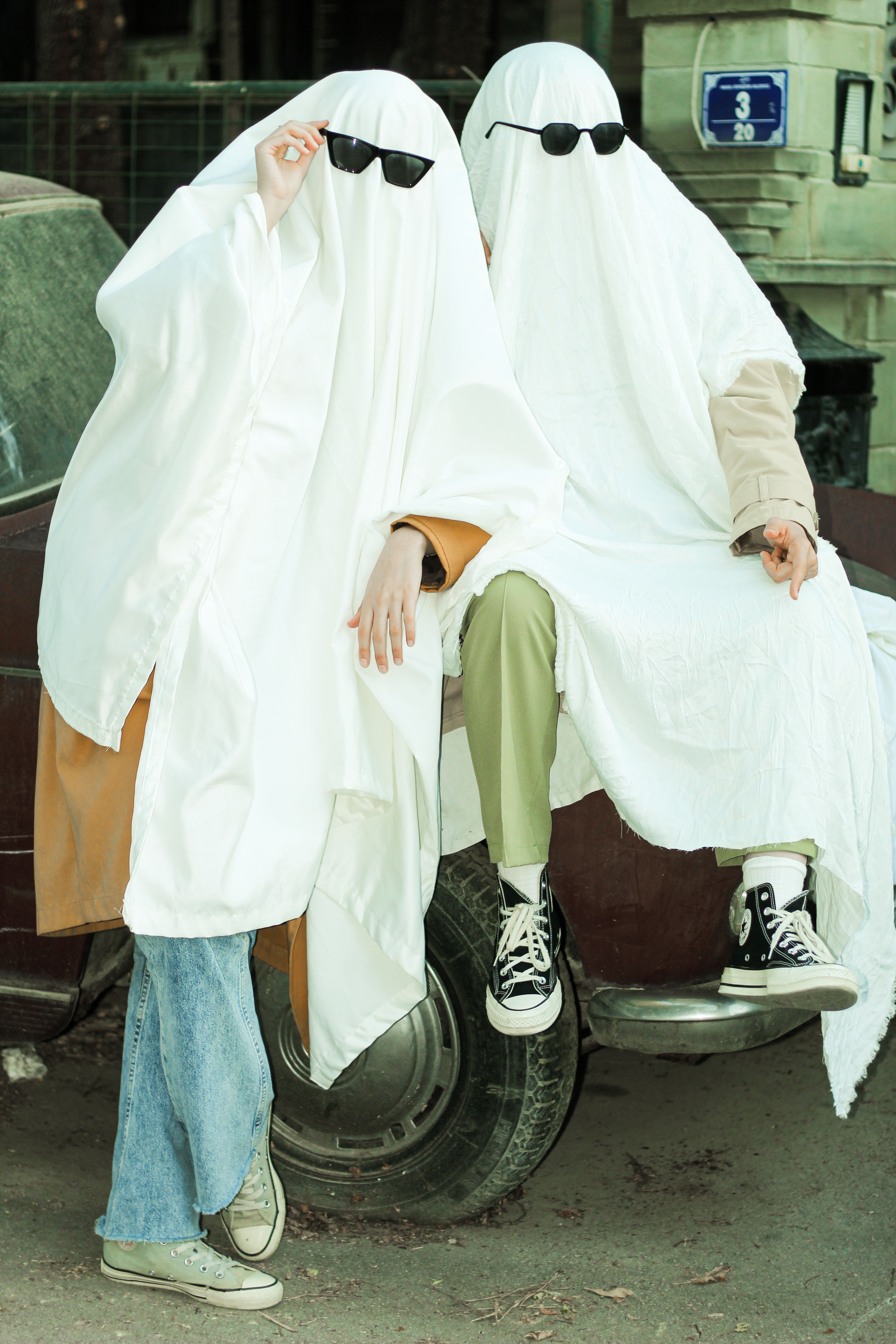 Two people dressed as ghosts with sunglasses sit on a car for Halloween fun.