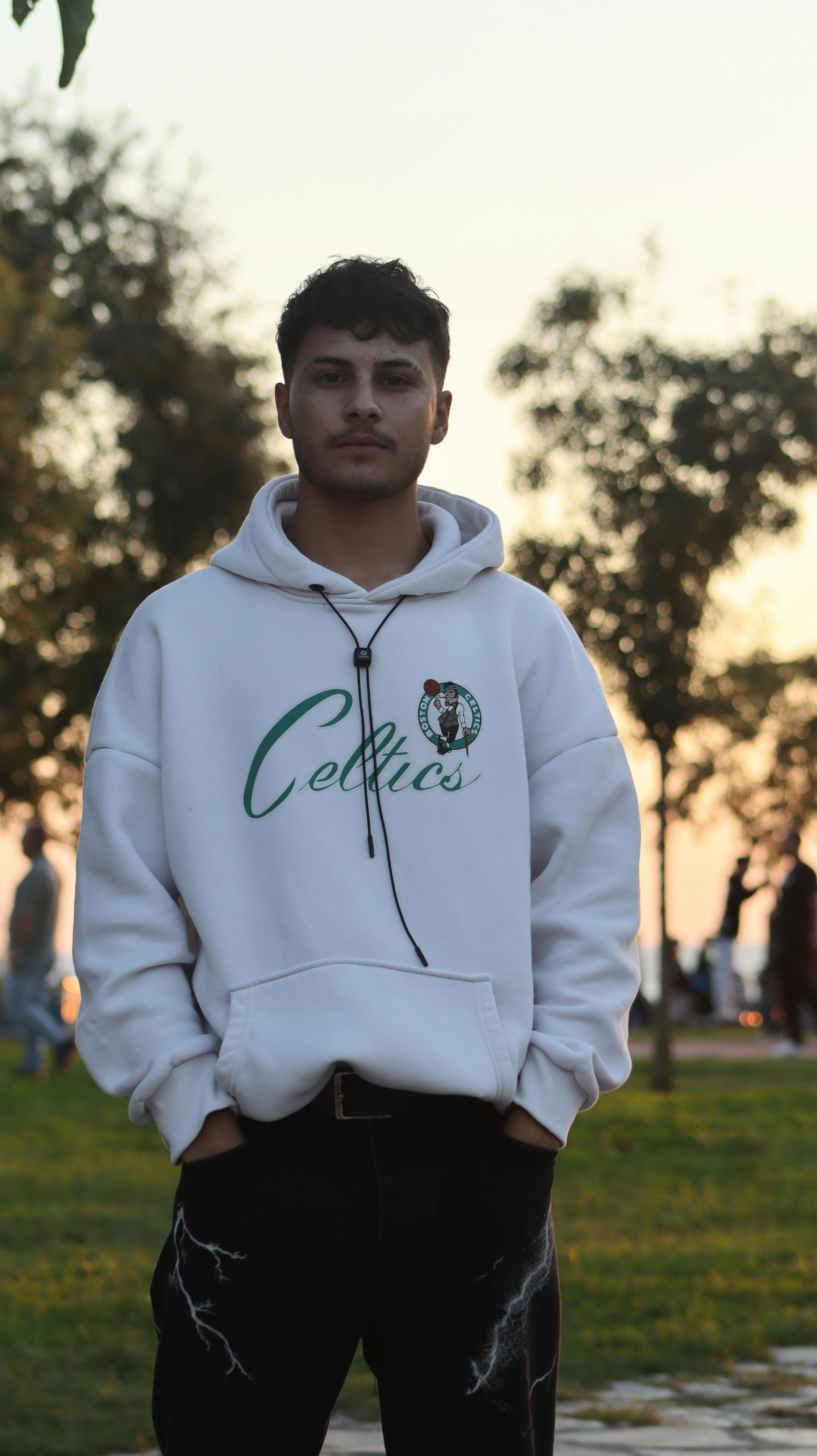 Young man wearing a hoodie standing outdoors at twilight with trees in the background.
