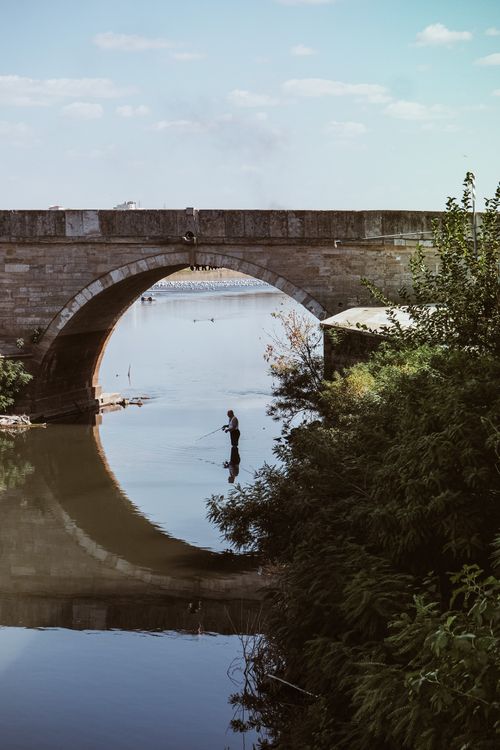 Free A serene scene of a person fishing under an old stone bridge arch, reflected in calm waters. Stock Photo