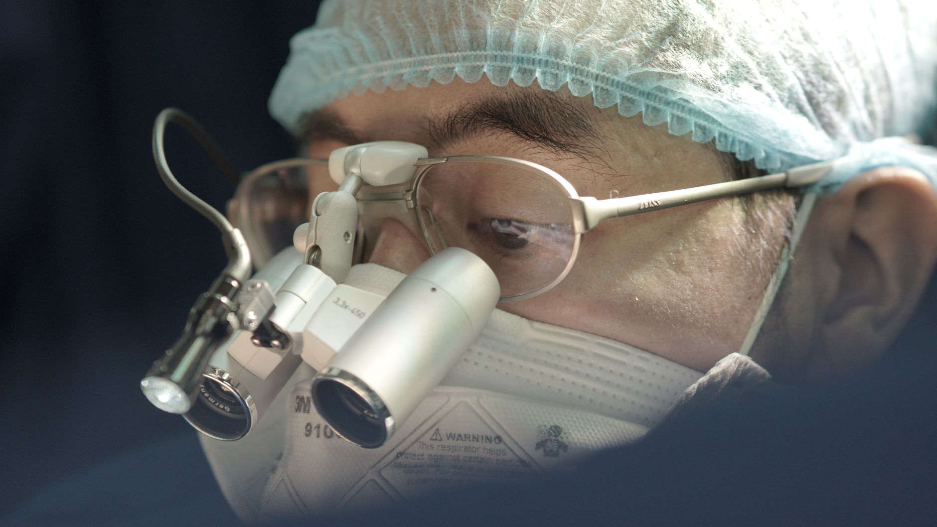 Focused surgeon wearing a surgical mask and loupes during an operation.