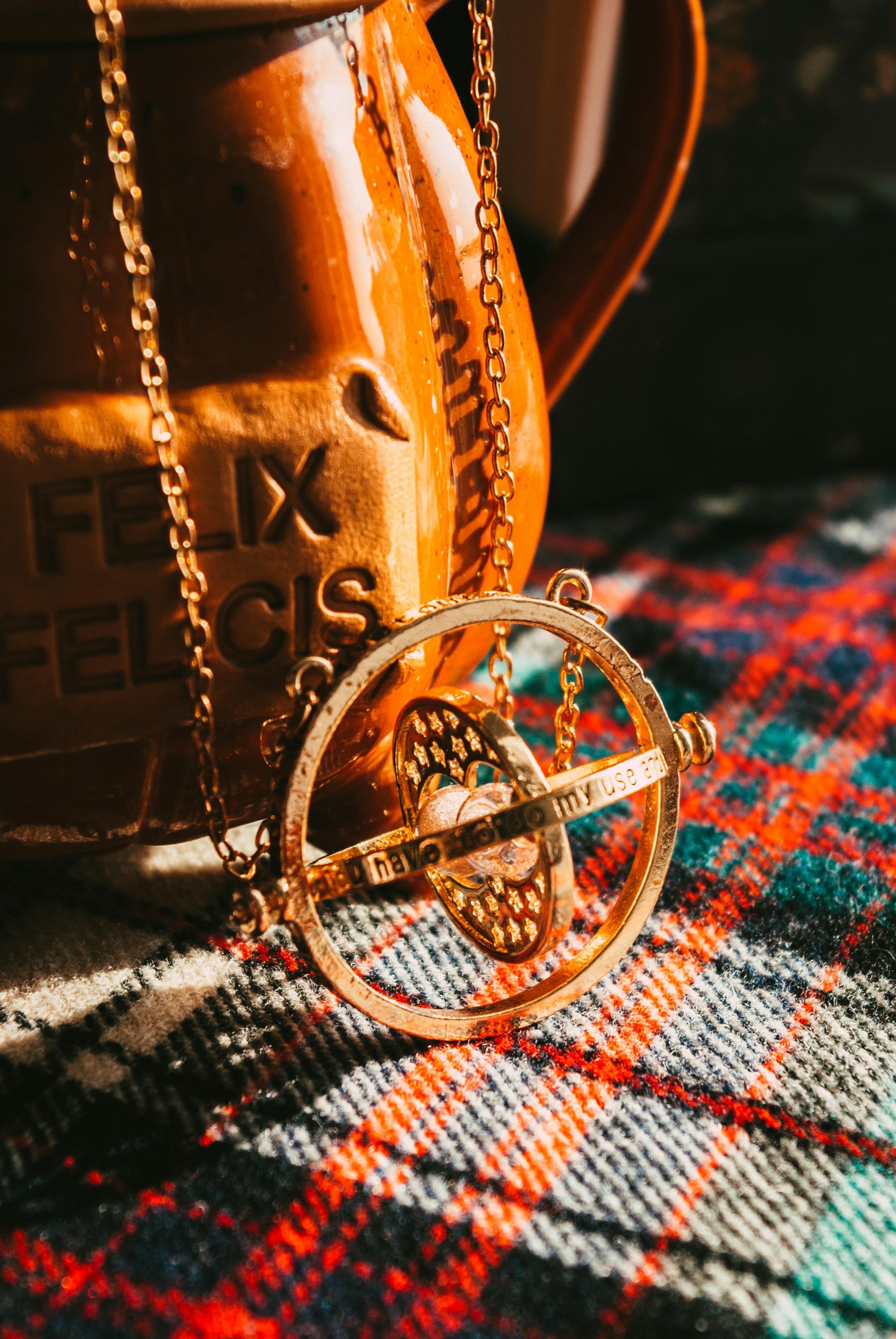 Close-up of a gold necklace with intricate design on a colorful tartan cloth beside a pottery mug.
