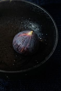 A single fresh fig sits in a black bowl with water droplets, highlighting its natural texture.
