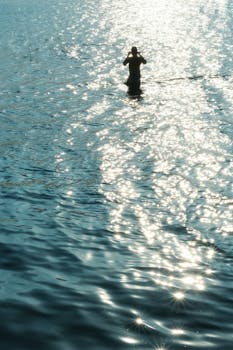 A silhouette of a person stands in the shimmering ocean waters at sunset, creating a serene atmosphere.