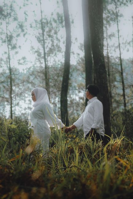 A couple elegantly dressed in white walking hand-in-hand through a lush forest, symbolizing love and togetherness.