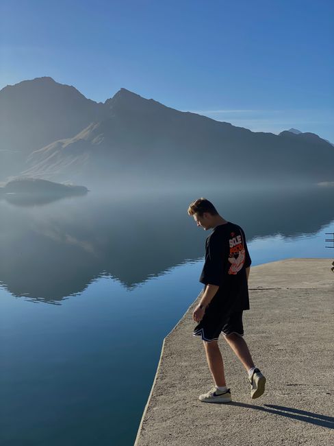 Young man on concrete pier gazing at serene lake and mountain reflections in Valloire, French Alps.
