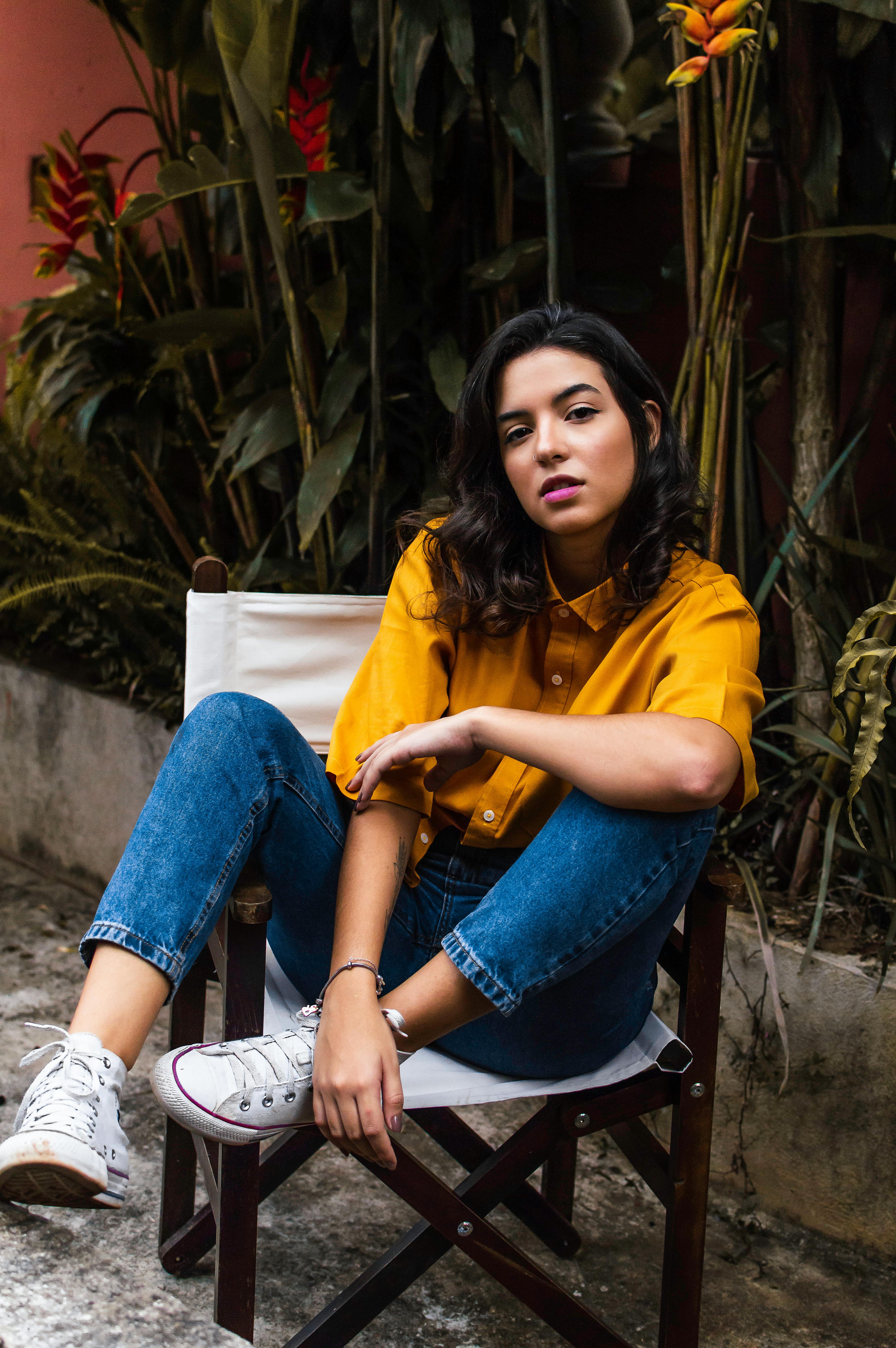 一位穿着黄色衬衫和牛仔裤的自信女人，坐在热带植物环绕的椅子上