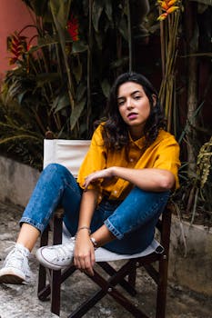 Confident woman in yellow shirt and jeans sitting on a chair, surrounded by tropical plants.