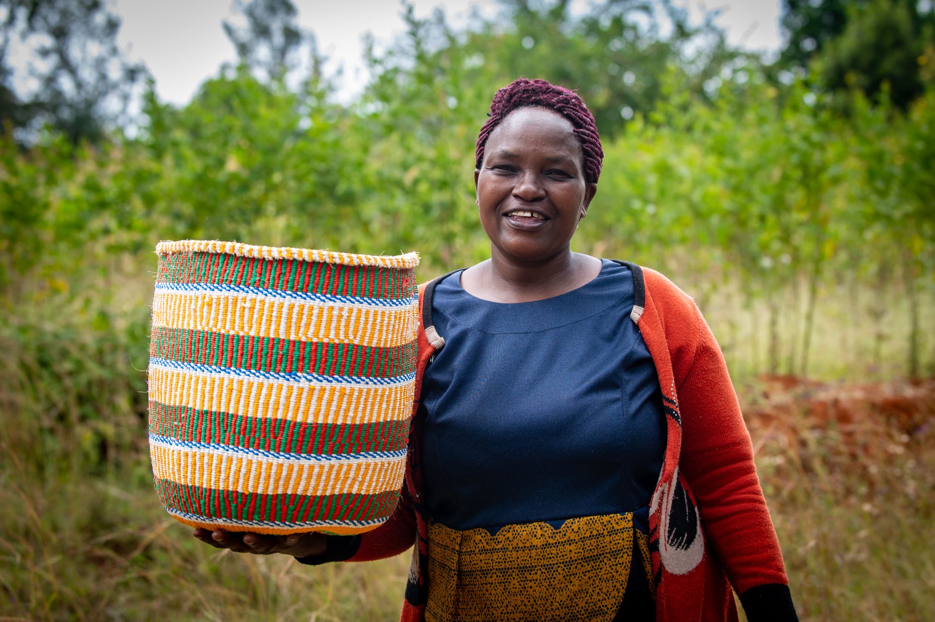 Smiling African woman in rural setting holding a colorful traditional basket, showcasing craftsmanship.