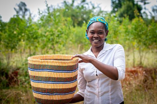 A smiling woman in a rural setting holds a colorful woven basket made by hand.