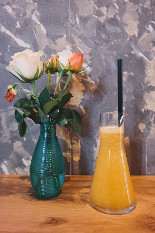 Free Clear Glass Pitcher With Yellow Mixture Near Green Glass Vase With Flowers on Table Stock Photo