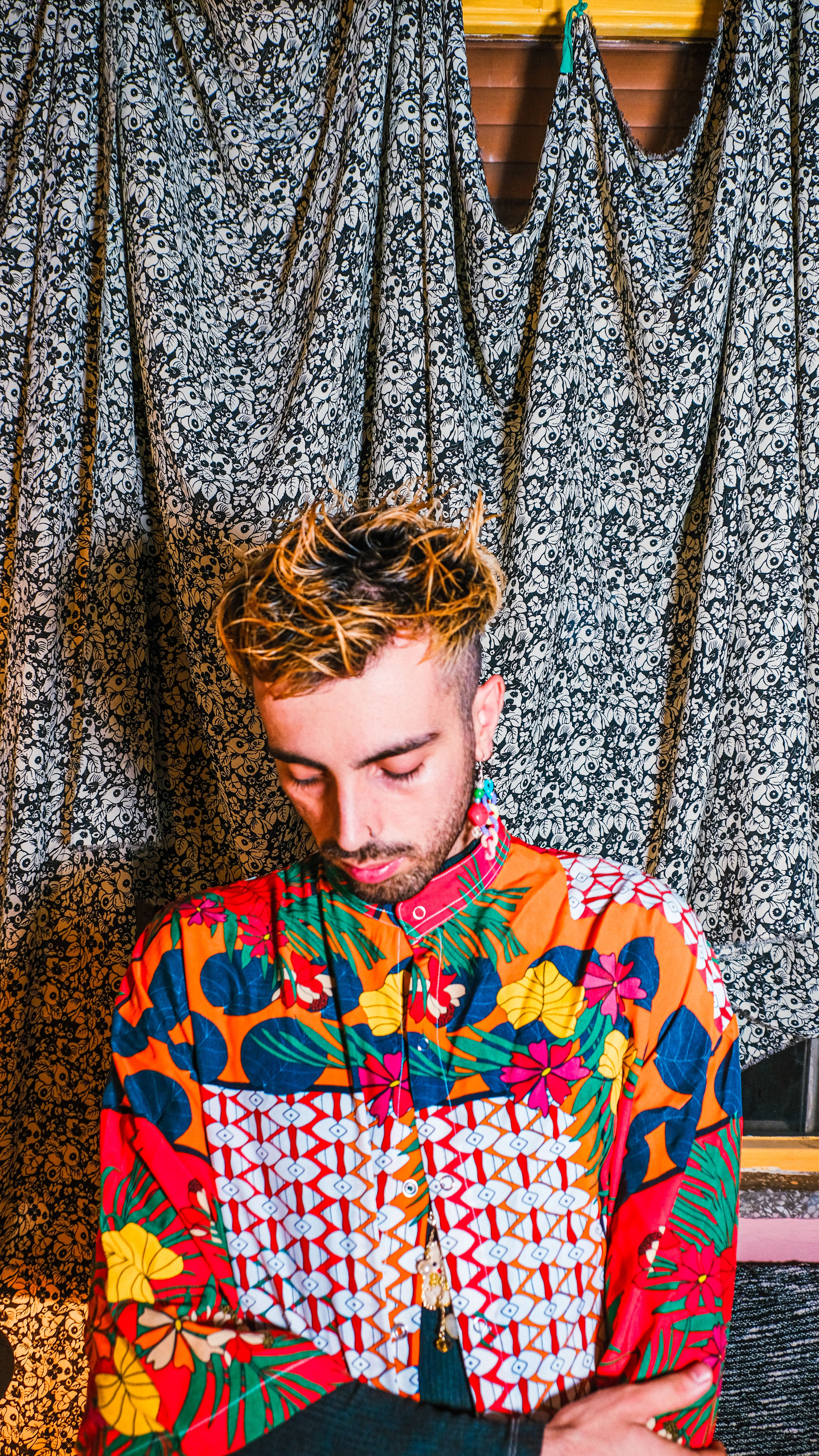 A male model wearing a colorful shirt, looking down, with a patterned background.