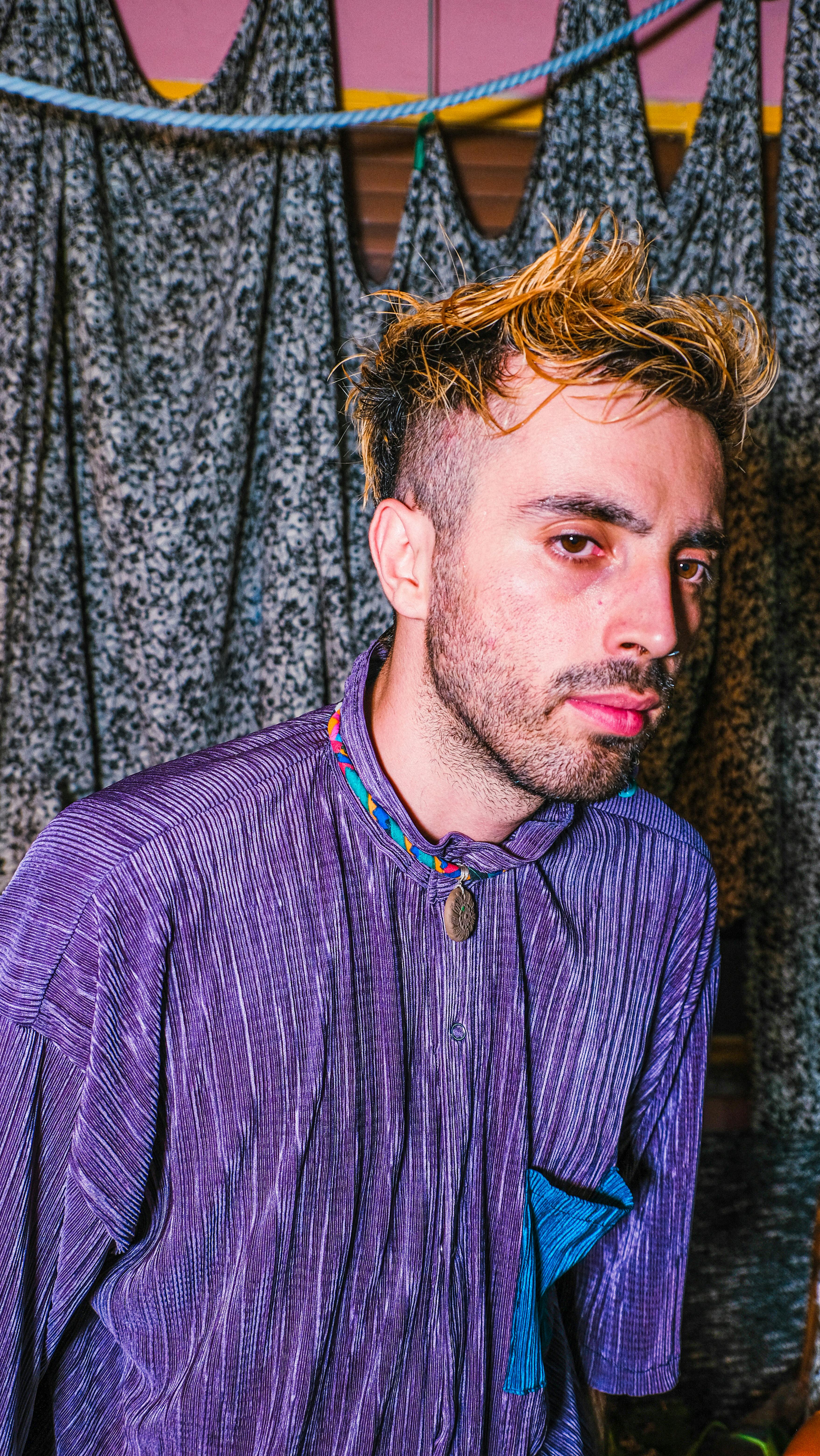 A stylish man with dyed short hair wearing a purple shirt in a fashion-forward pose.