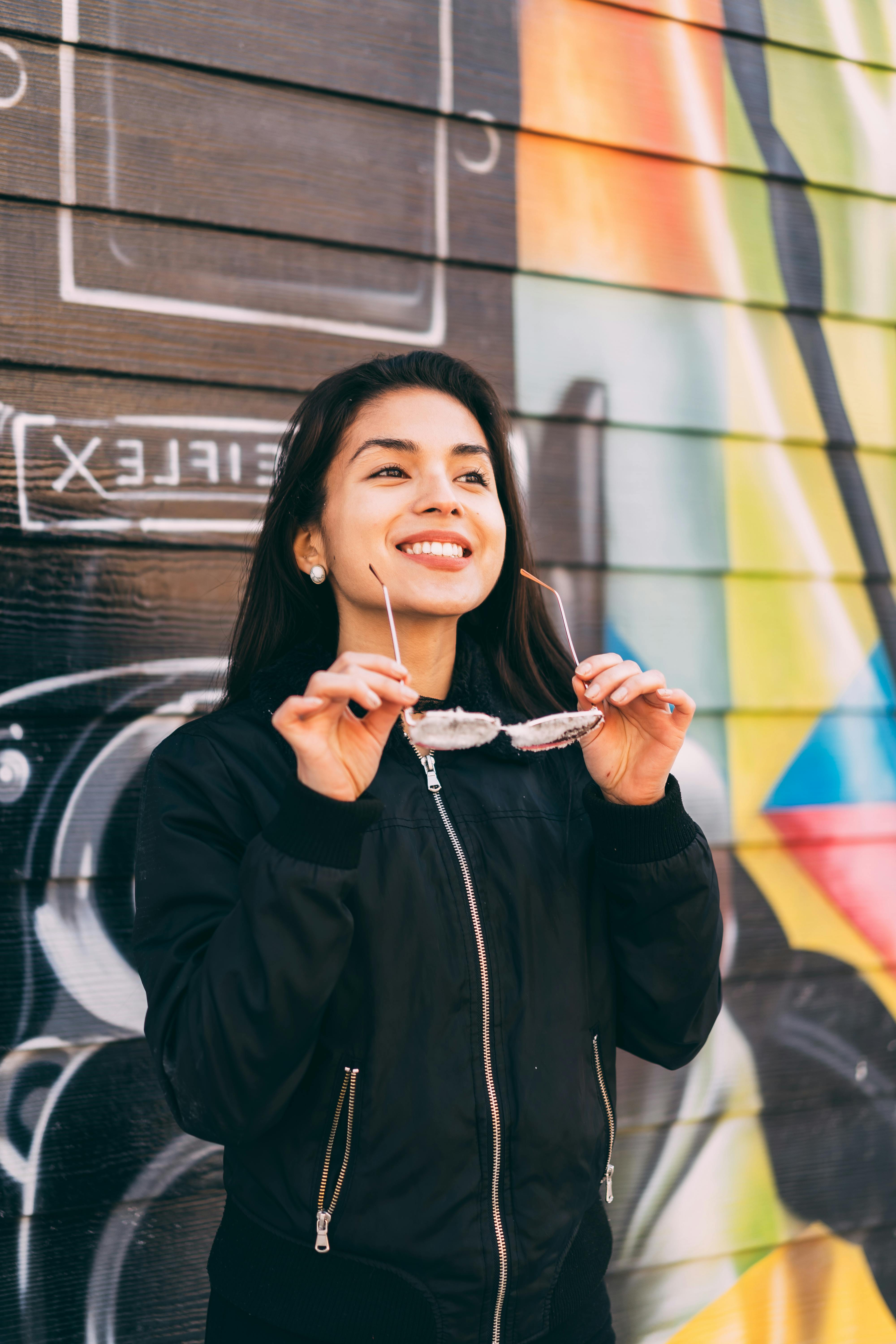 一位面带微笑的女子，手持太阳镜，倚靠在色彩斑斓的涂鸦墙边，展现出喜悦与休闲的风格