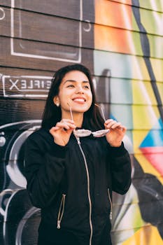 一位面带微笑的女子，手持太阳镜，倚靠在色彩斑斓的涂鸦墙边，展现出喜悦与休闲的风格