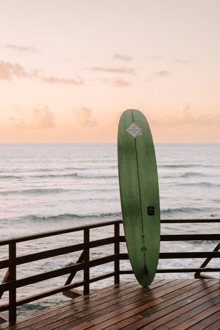 A green surfboard stands on a wooden deck overlooking the calm ocean waves at sunset.