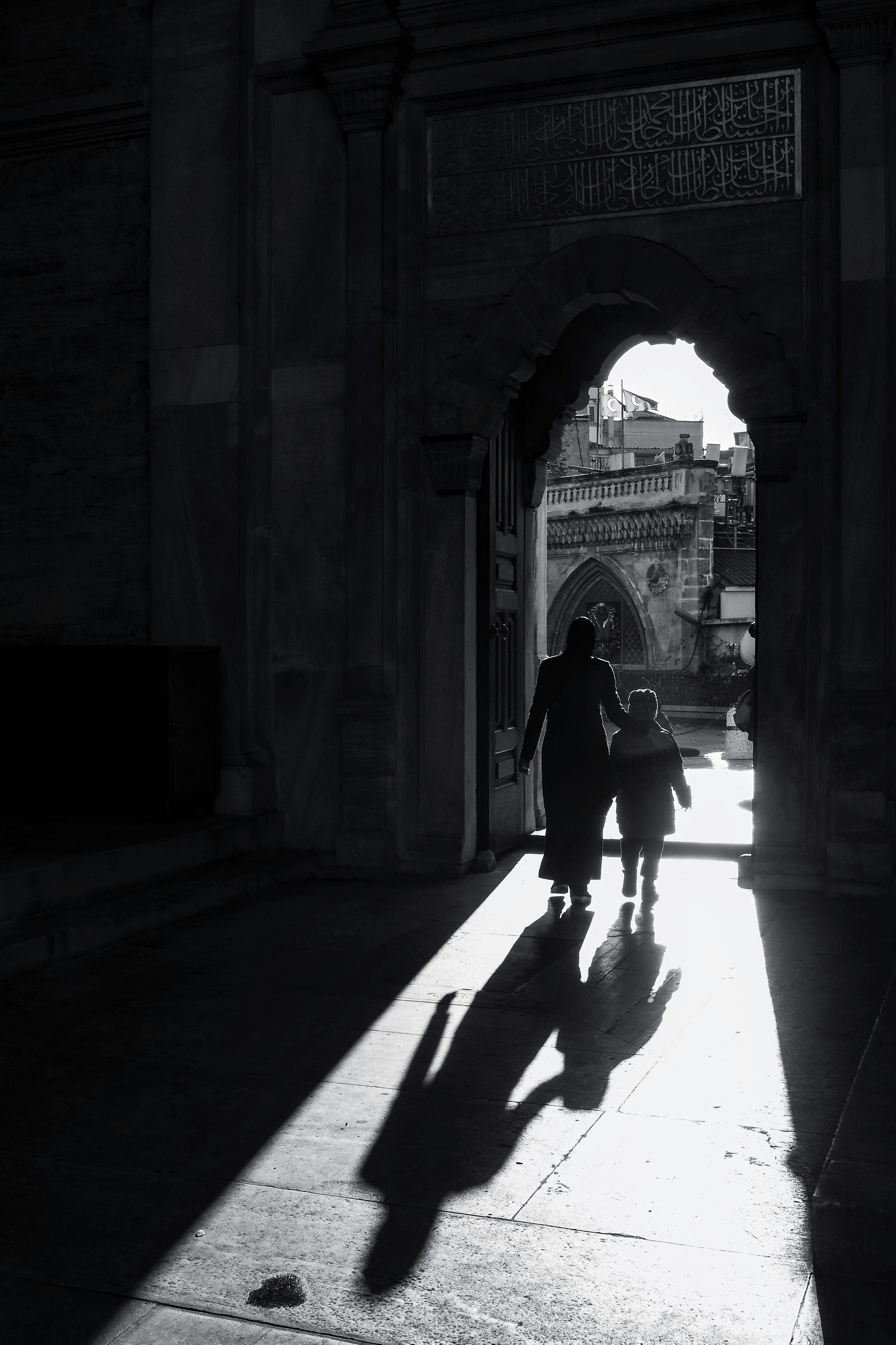 Silhouette of a mother and child walking through an archway of a mosque with sunlight casting shadows.