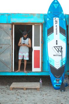 Man standing at a beach hut entrance with a paddleboard, ready for a surf adventure.