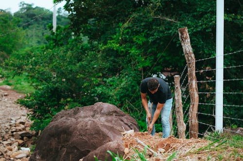 Man in T-Shirt Digging in Ground by Fence with Wooden Stick