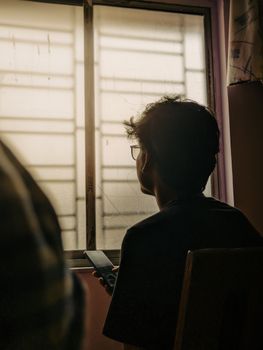 Silhouette of man with mobile phone, in sunlight through window, conveying solitude and reflection.