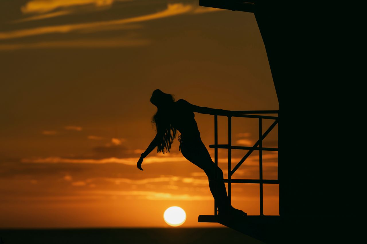 a woman backlit in silhouette leans over a balcony at sunset exuding f