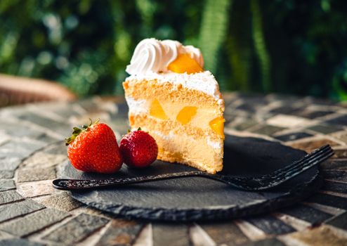 A delectable slice of fruit cake topped with cream and accompanied by fresh strawberries and a fork on a stone plate.