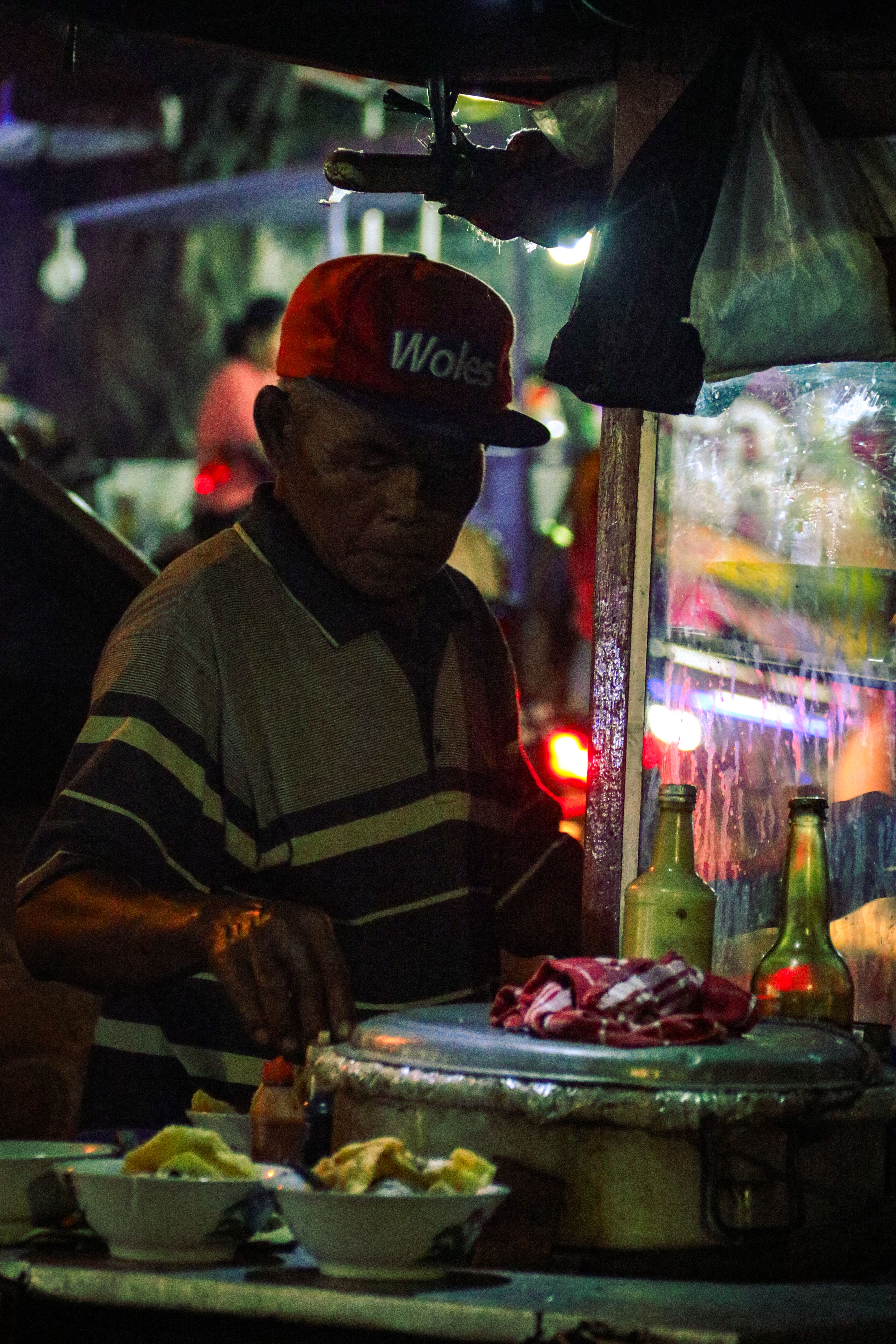 Elderly man serves street food at night with warm lighting and vibrant atmosphere.
