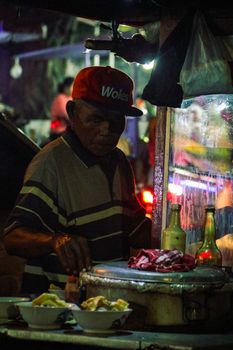 Elderly man serves street food at night with warm lighting and vibrant atmosphere.
