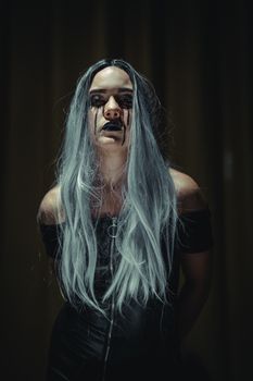 A gothic styled woman with dyed gray hair and dramatic makeup stands against a dark background.