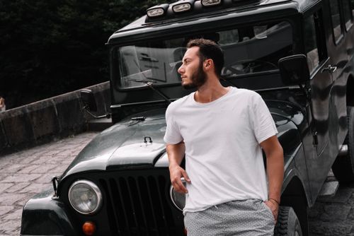 Free Portrait of a young man in a t-shirt leaning on a classic SUV in an outdoor setting. Stock Photo