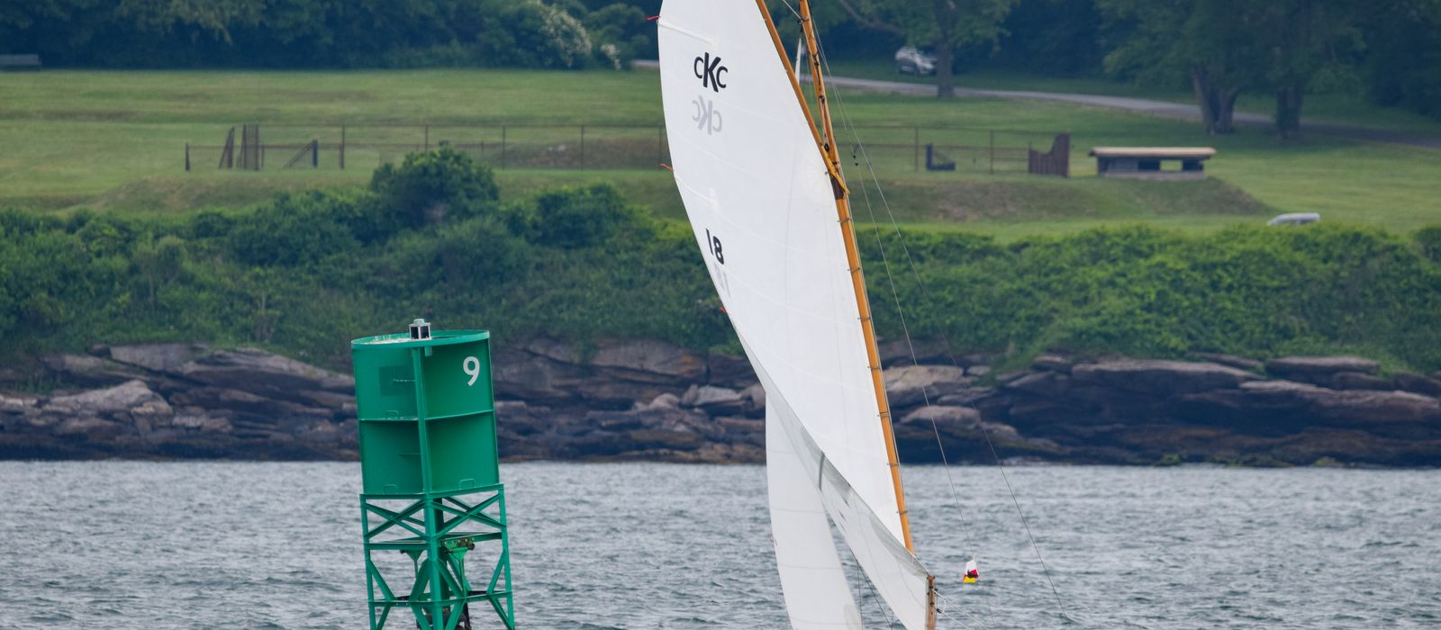 Sailboat navigating near a green buoy in Newport, RI, against a lush landscape.