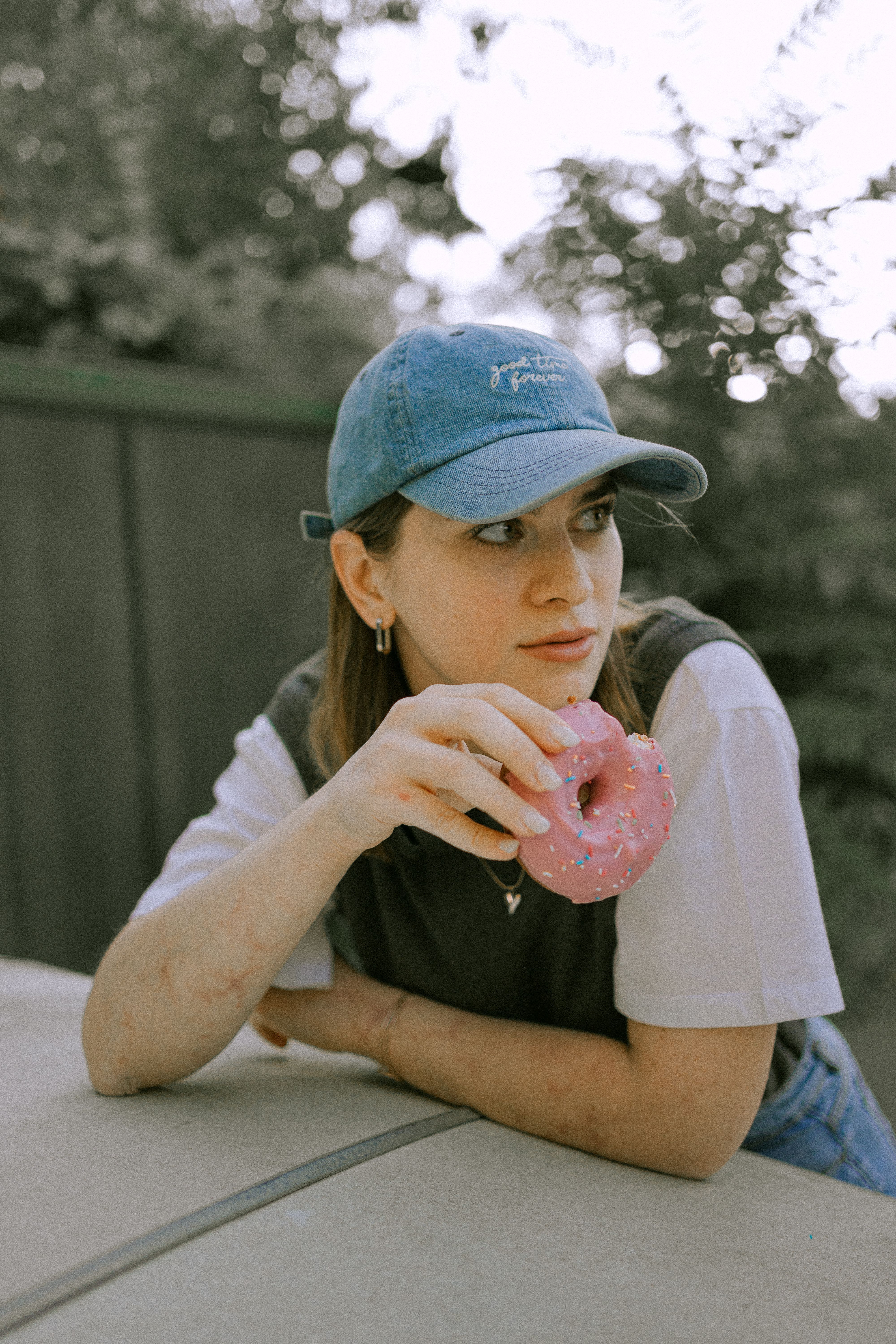 Young woman leans on table, holding a pink donut. Casual outdoor portrait.