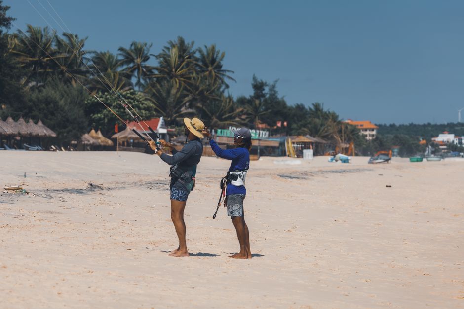 Two people learning kitesurfing on a sunny tropical beach with palm trees.