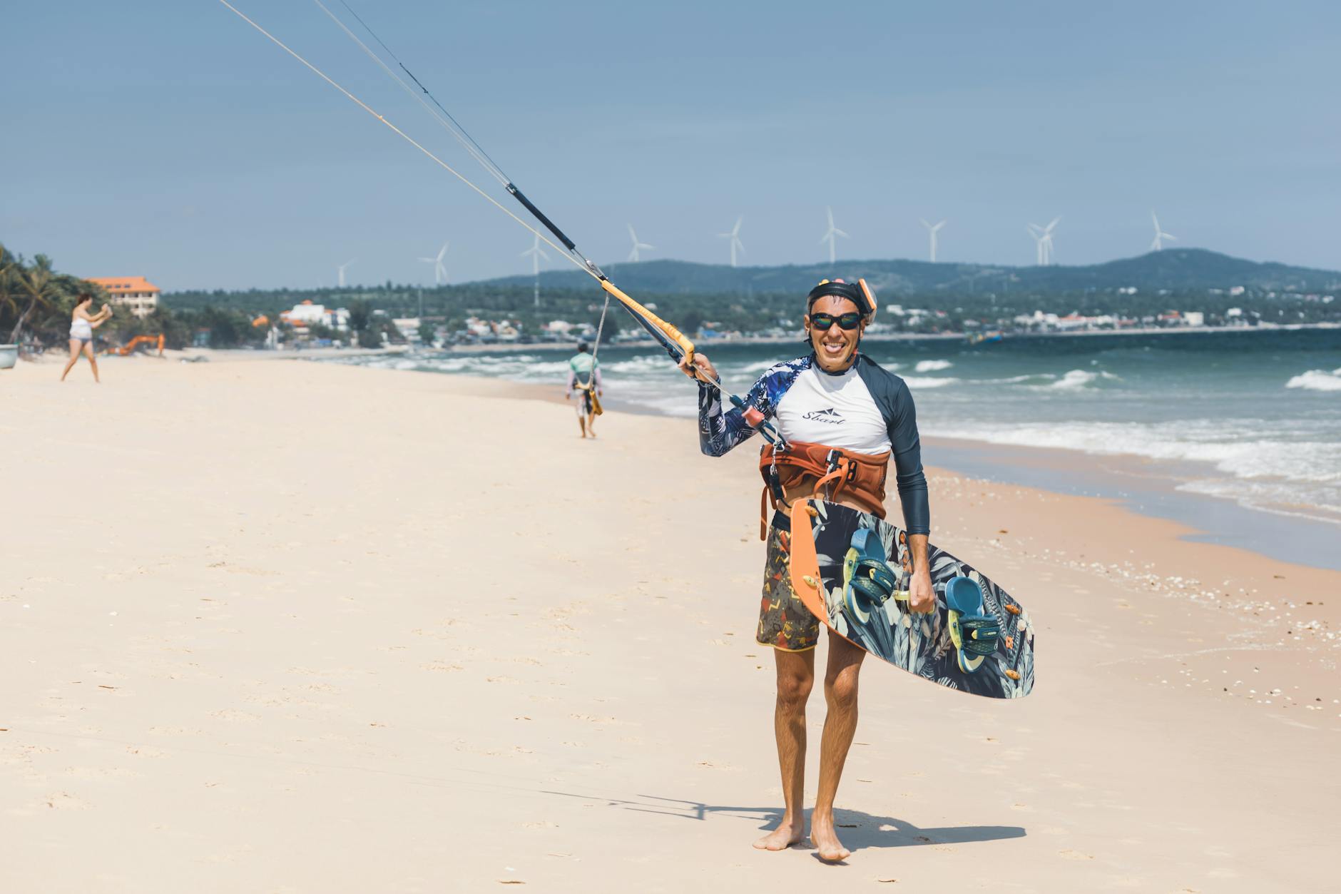 Smiling kitesurfer holding board on a sunny beach with wind turbines in the background.