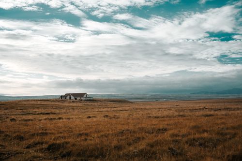 Free A solitary abandoned house stands amidst expansive rural grasslands under a cloudy blue sky. Stock Photo