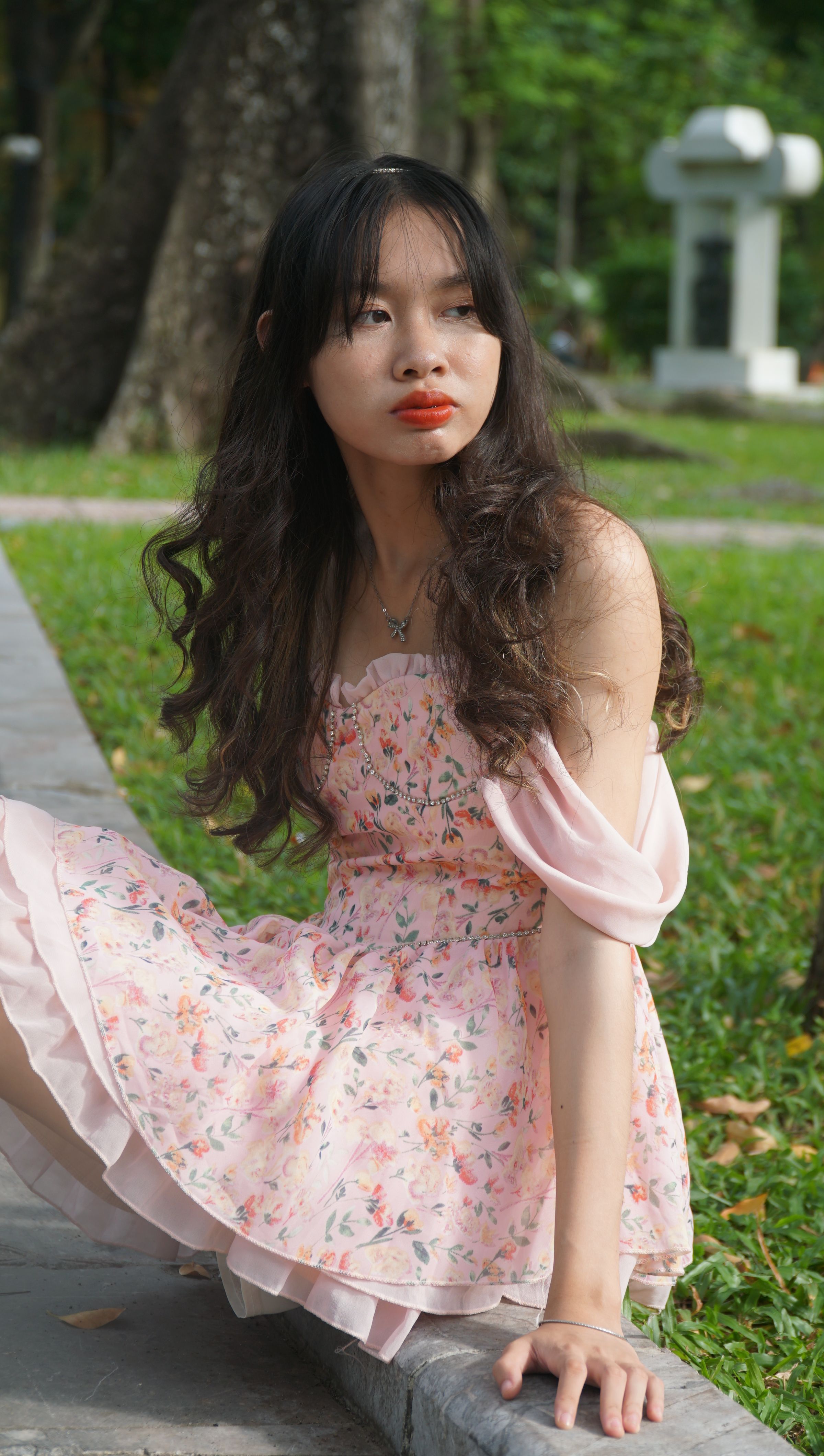 A young woman in a floral pink dress poses gracefully in an outdoor park setting.