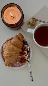 Top view of a croissant and fig slices with tea and candle on a white table.